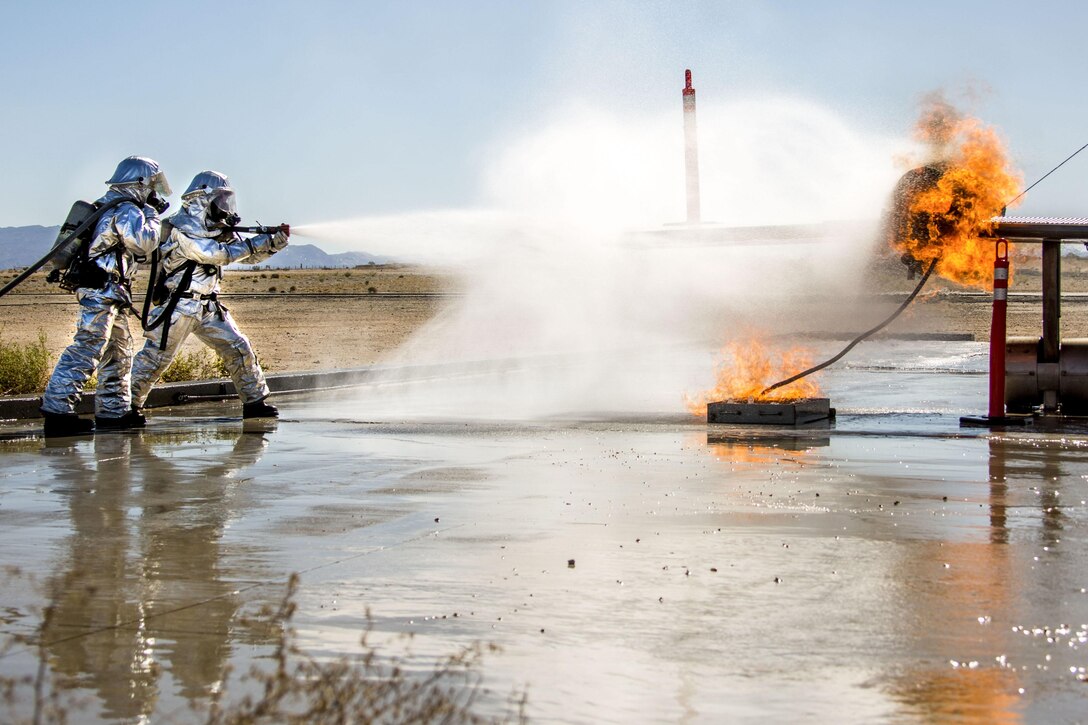 Two Marines in silver protective garb spray water from a hose onto a blaze.