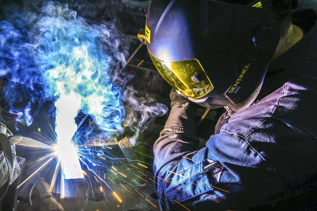 A sailor wearing a helmet does welding work, creating sparks and blue smoke.