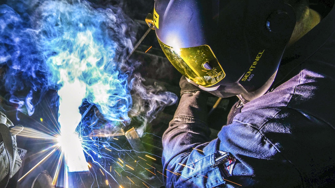 A sailor wearing a helmet does welding work, creating sparks and blue smoke.