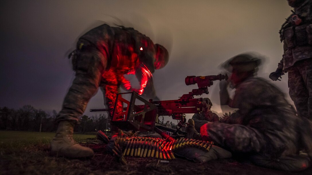 A soldier, illuminated by red light against a dark sky, preps a machine gun as another looks through a scope on it.