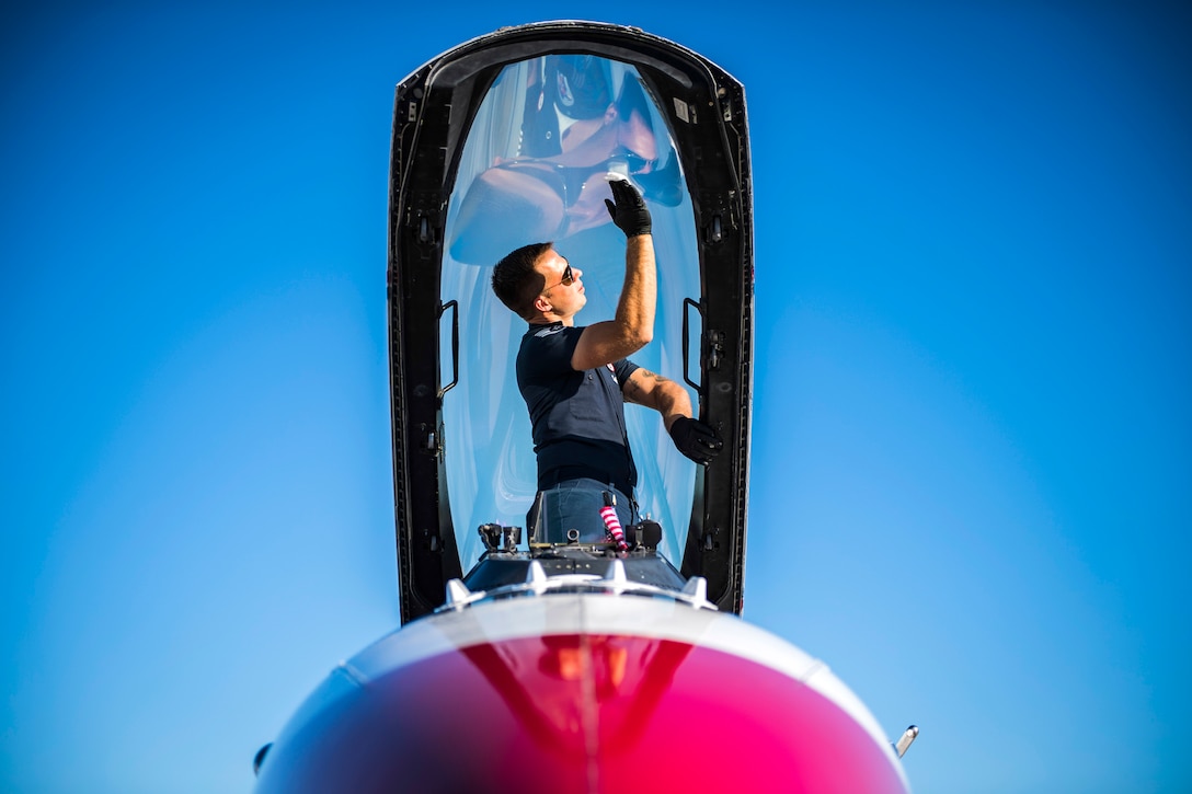 An airman stands in a fighter jet's open cockpit and wipes the glass canopy.