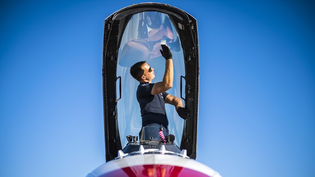 An airman stands in a fighter jet's open cockpit and wipes the glass canopy.