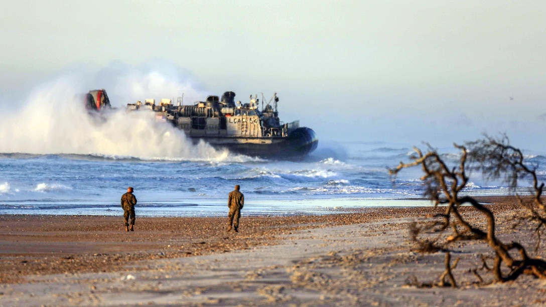 A landing craft creates large wake in the water while heading to shore, where service members are walking.