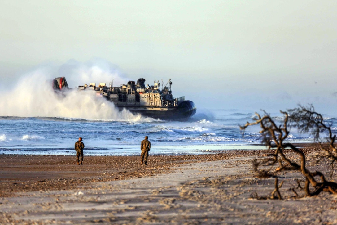 A landing craft creates large wake in the water while heading to shore, where service members are walking.