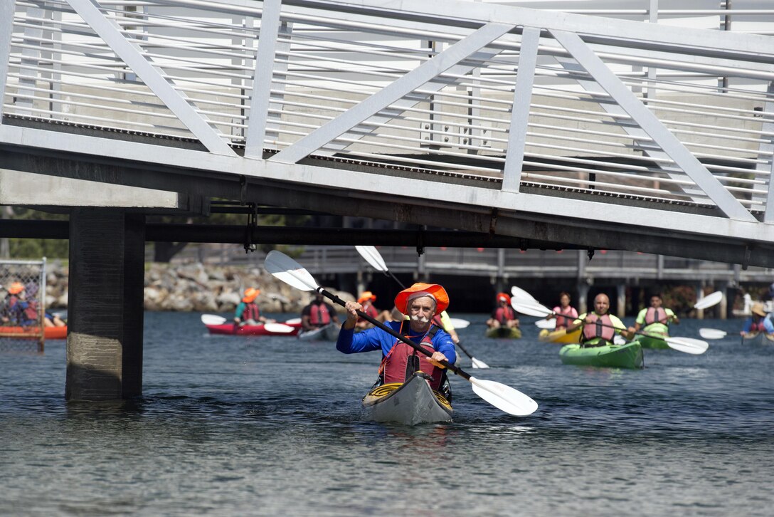 A group of patients kayak on a bay below a walkway overpass.