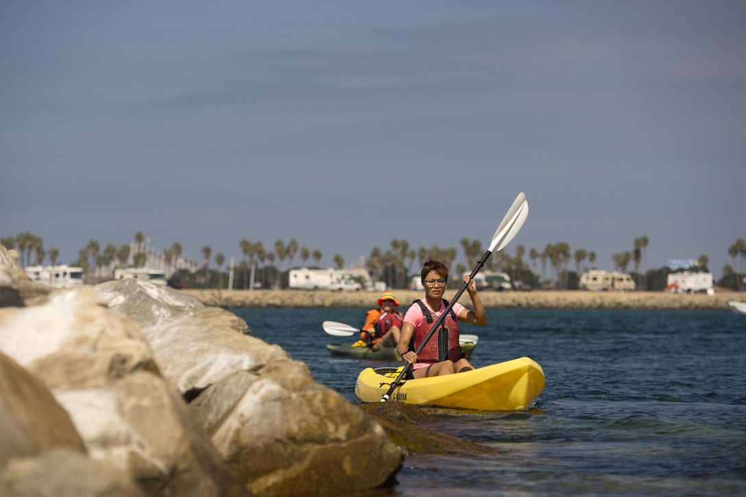 A kayaker paddles near rocks on a bay with another behind her.