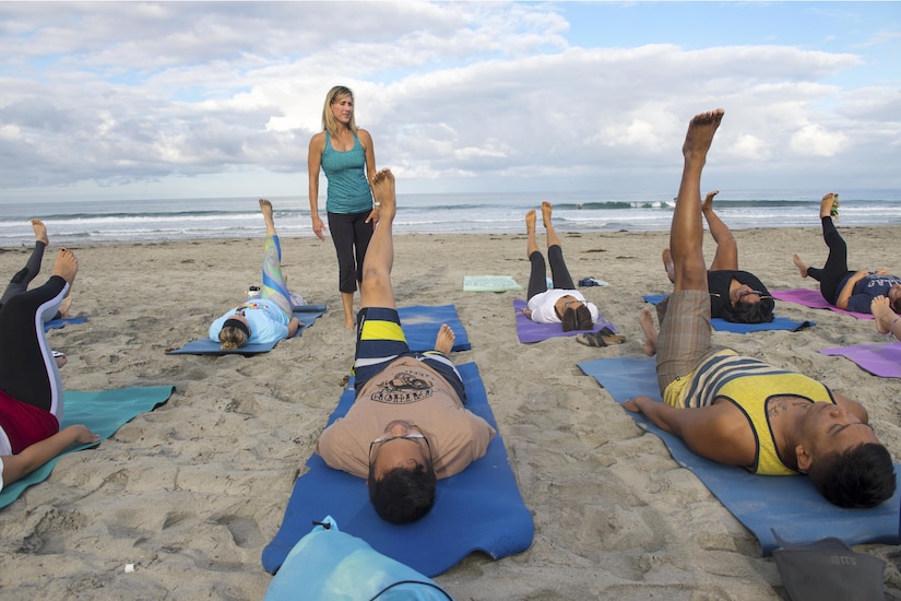 An instructor stands while a group of yoga participants lay on the beach with one leg up.