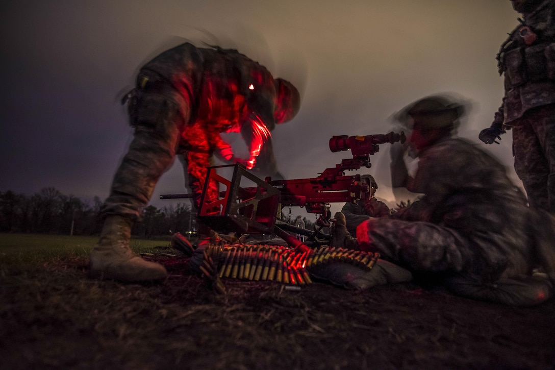 A soldier, illuminated by red light against a dark sky, preps a machine gun as another looks through a scope on it.