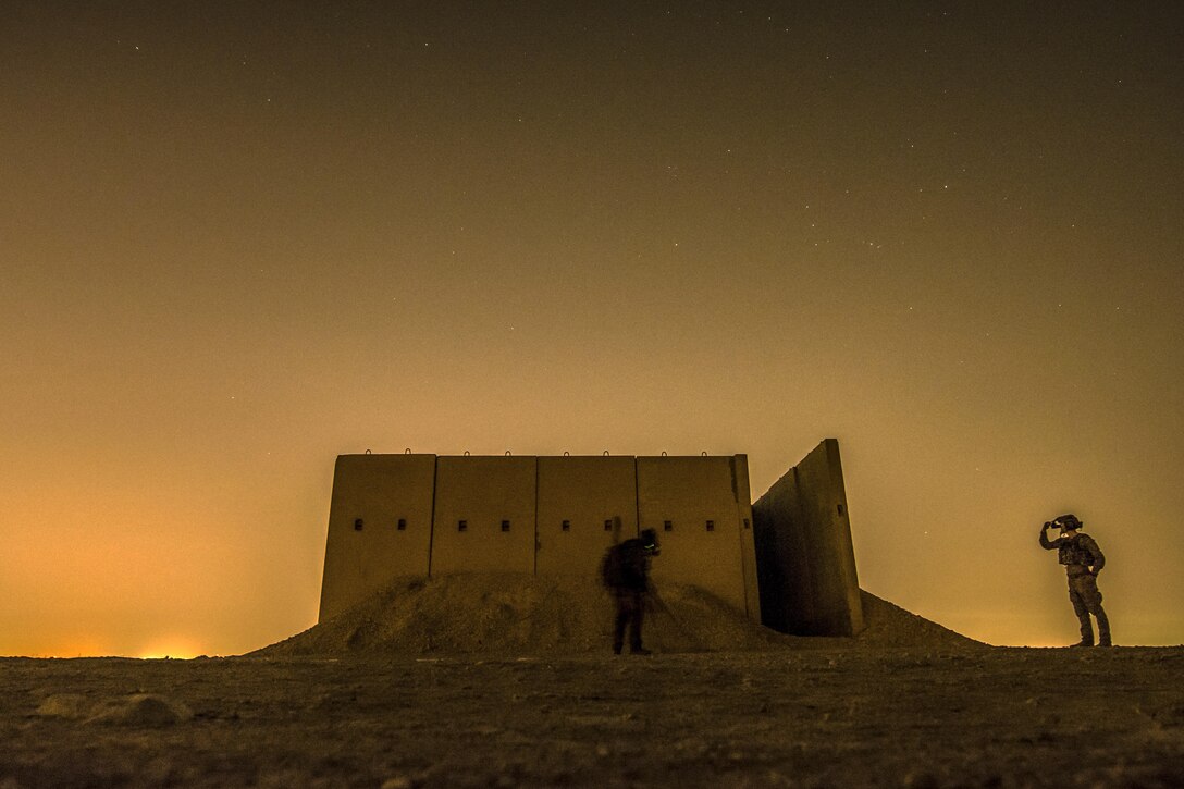 An airman uses a metal detector by a structure as another one watches, against a darkening sky.