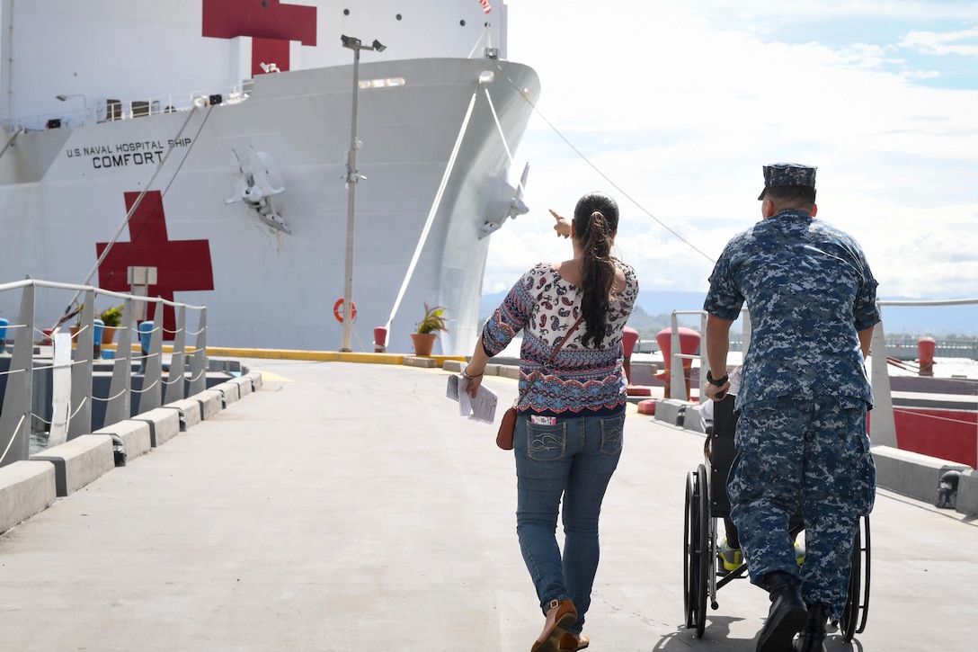 A sailor pushes a wheelchair and walks with a person to a ship.