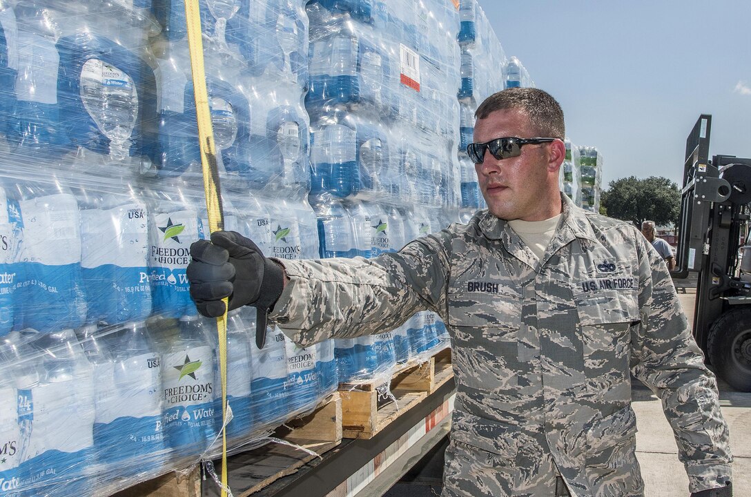 Soldier in fatigues secures pallet of water with tie-down strap, facing viewer's half-left.