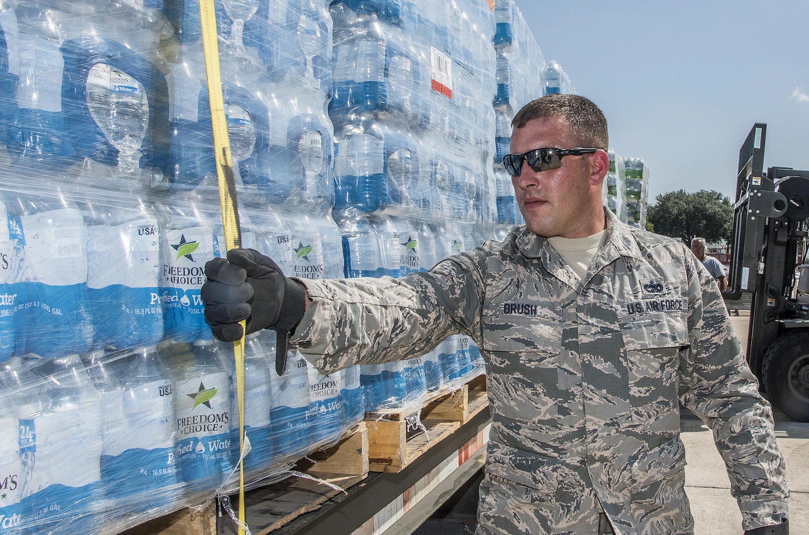 Soldier in fatigues secures pallet of water with tie-down strap, facing viewer's half-left.