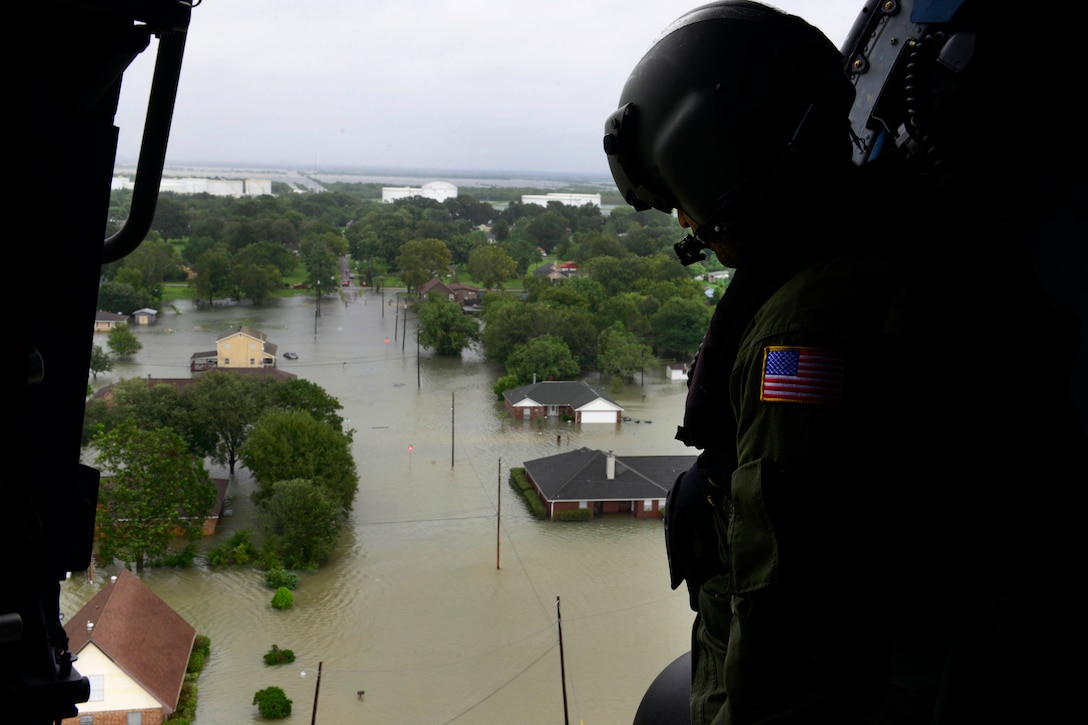 A Coast Guardsman peers out of the door of an aircraft flying over flooded streets.