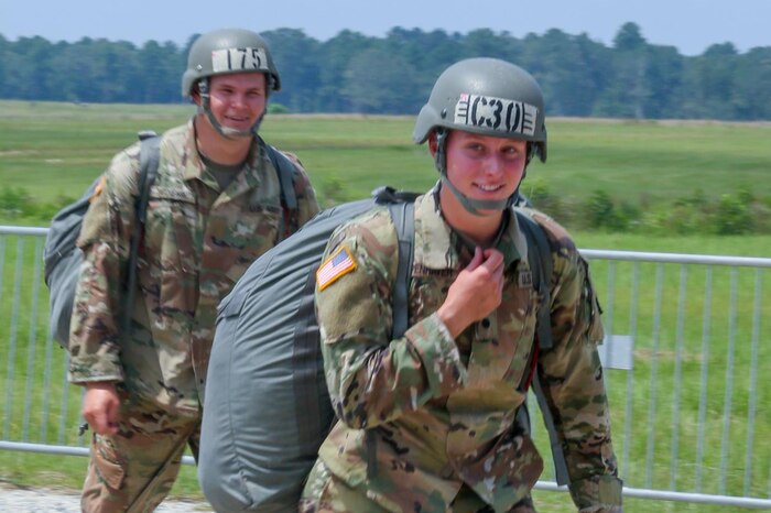 Two soldiers walk with parachute gear on their backs.