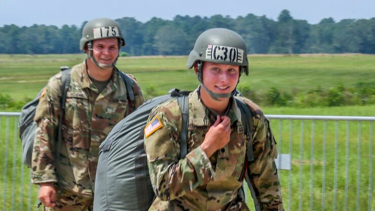 Two paratroopers walk with their gear across a grassy drop zone.