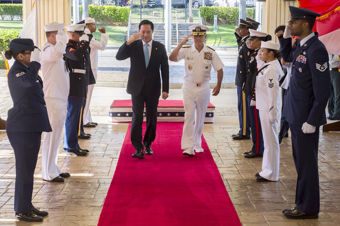 Two people salute while walking down a red carpet with American service members lined on each side.