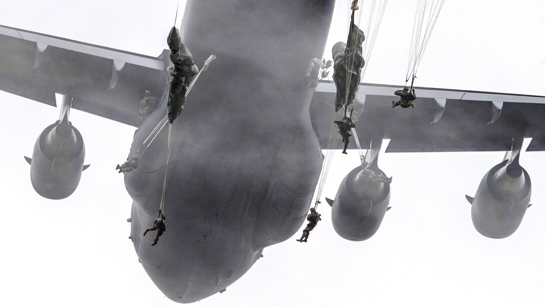 Soldiers bodies and parachutes frame an aircraft as they jump from it.