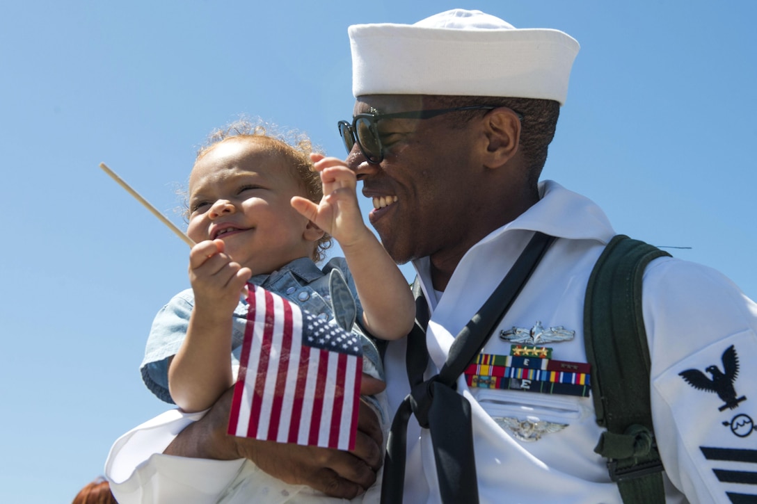 A sailor smiles as he holds his baby daughter, who is smiling and holding a small U.S. flag.