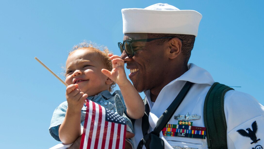 A sailor smiles as he holds his baby daughter, who is smiling and holding a small U.S. flag.