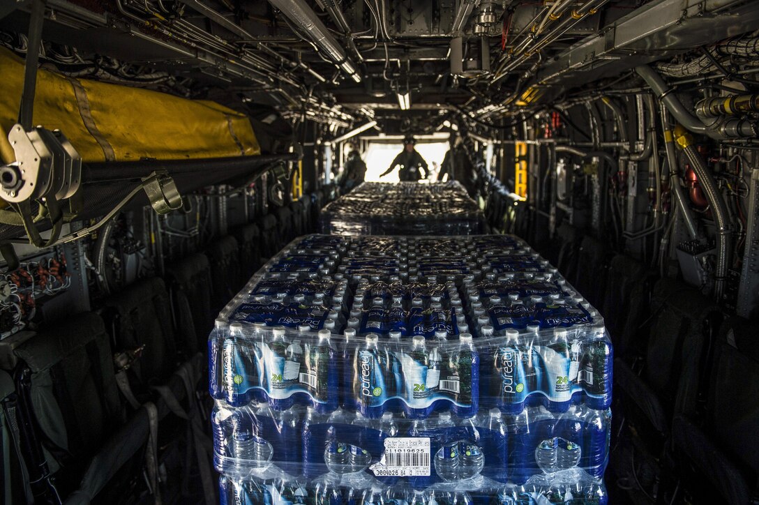 A pallet of water sits on a helicopter while service members work in the background.