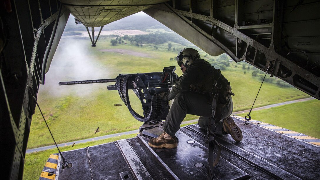 A Marine fires a machine gun from the back of a helicopter flying over green terrain.