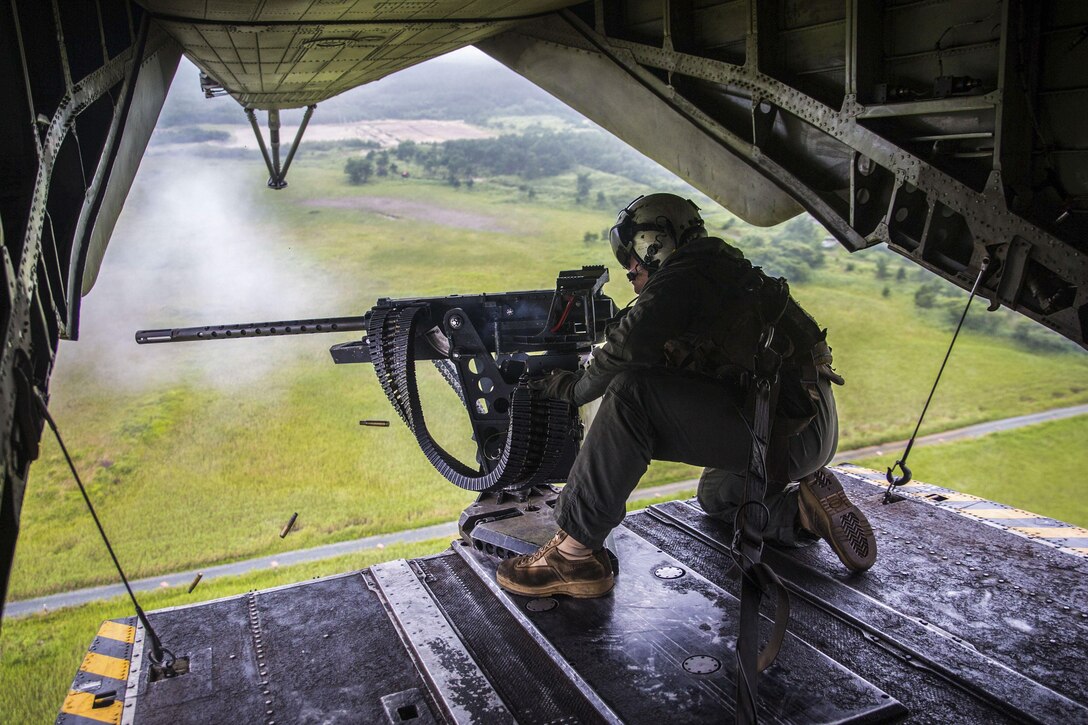 A Marine fires a machine gun from the back of a helicopter flying over green terrain.