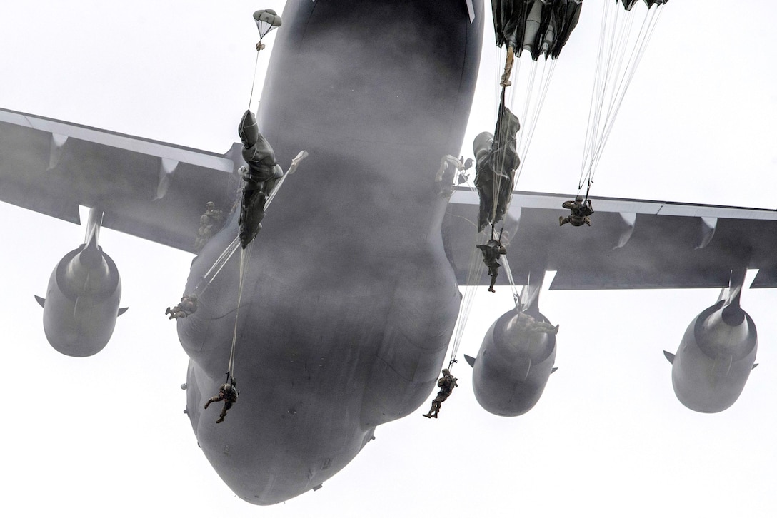 Soldiers bodies and parachutes frame an aircraft as they jump from it.