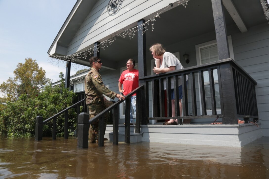 A soldier stands on the steps of a flooded house talking to two residents on the porch.