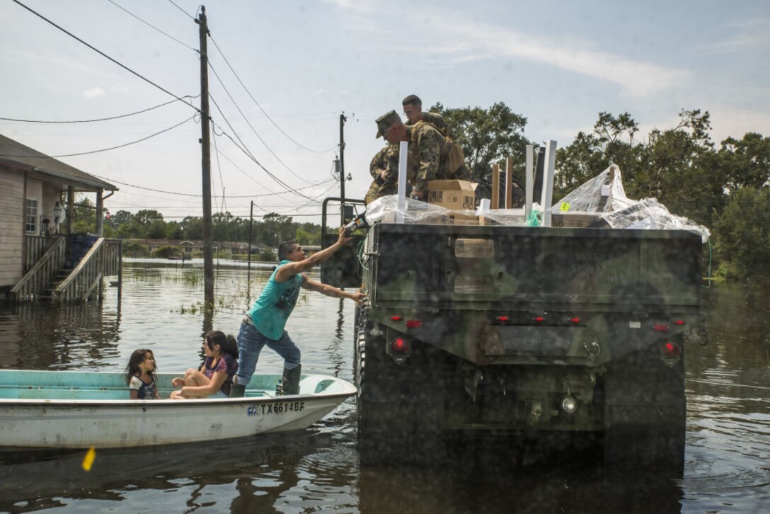 Marines on a military vehicle hand supplies to a man standing on a boat that is also carrying two young girls.