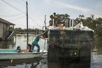 Marines on a military vehicle hand supplies to a man standing on a boat that is also carrying two young girls.