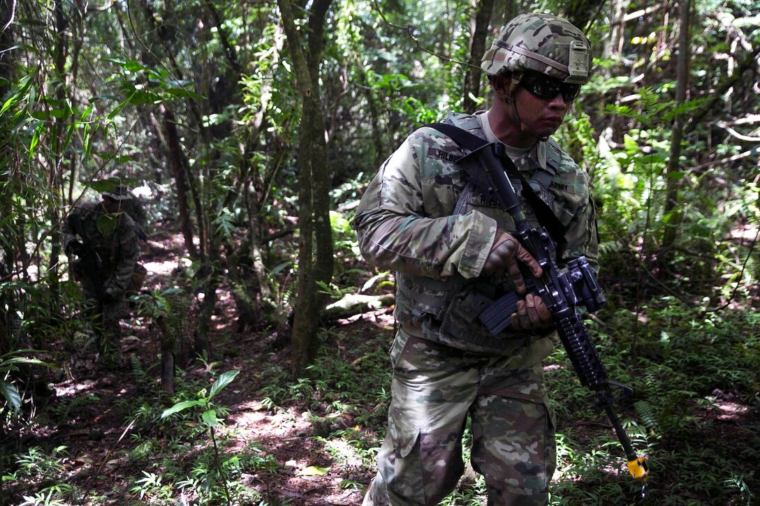 A soldier and Marine walk though a forested area with guns.