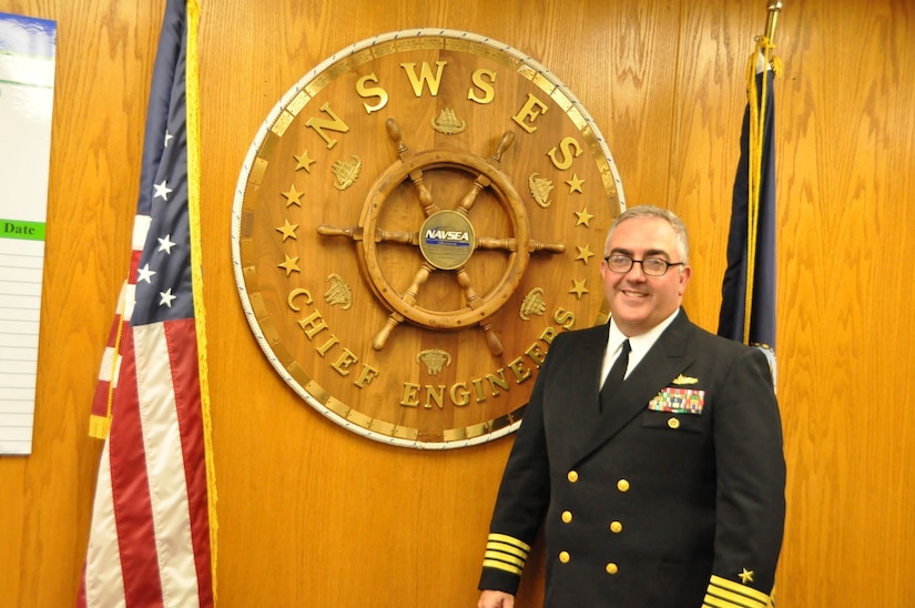 Navy Capt. Michael Ladner, chief engineer for Naval Surface Warfare Center, Port Hueneme Division in California, stands next to the wheel-shaped plaque that bears the names of all warfare center division chief engineers who served before him, July 20, 2017. Navy photo by Brian Melanephy