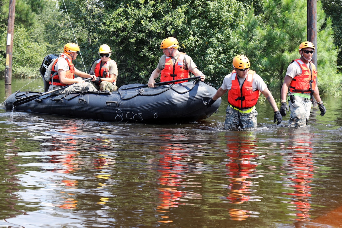A group of guardsmen wade through floodwaters pulling a boat with three other guardsmen.