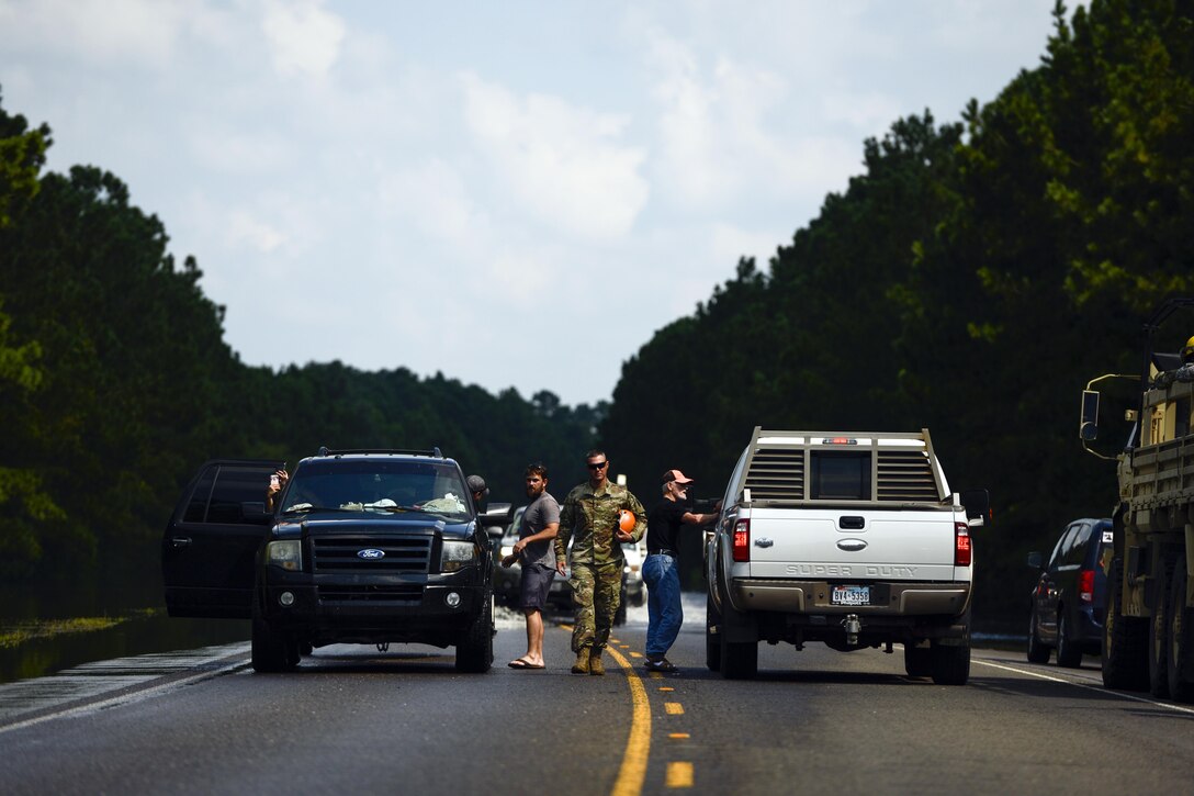A group of people and a soldier stand next to pickup trucks and a military vehicle.