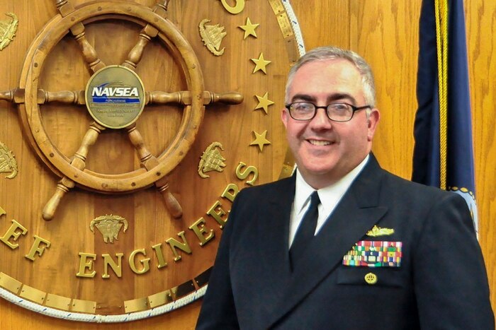 A sailor poses for a photo in front of an engineering symbol on a wood wall.