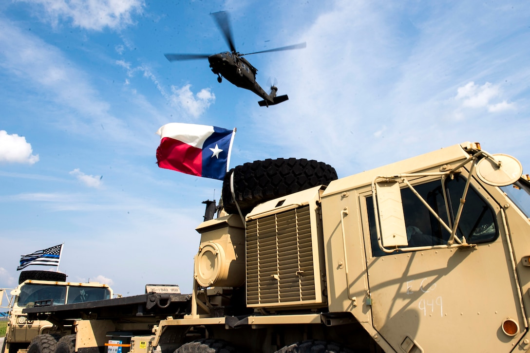 A helicopter flies in the air above military vehicles with a Texas flag.
