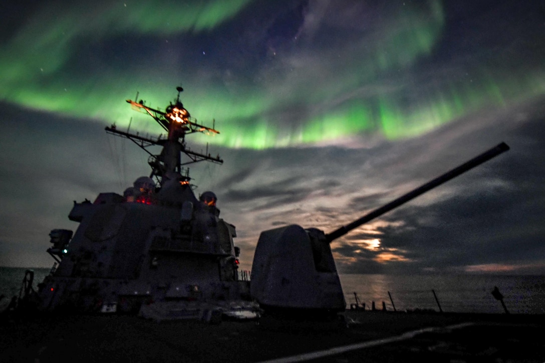 A destroyer in silhouette transits the ocean as northern lights flash green in the night sky.
