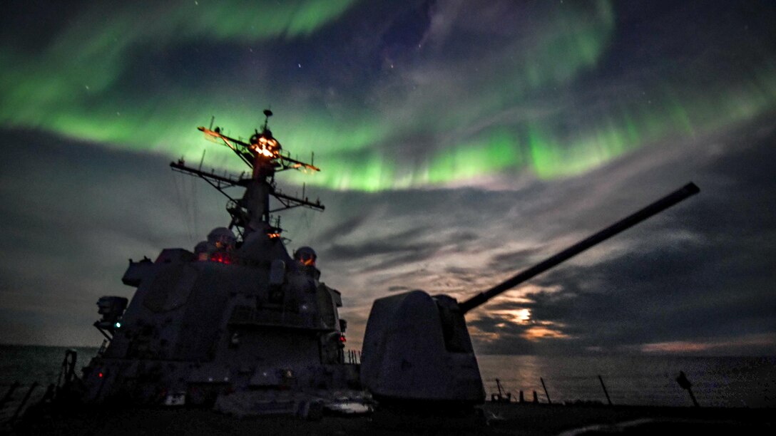 A destroyer in silhouette transits the ocean as northern lights flash green in the night sky.