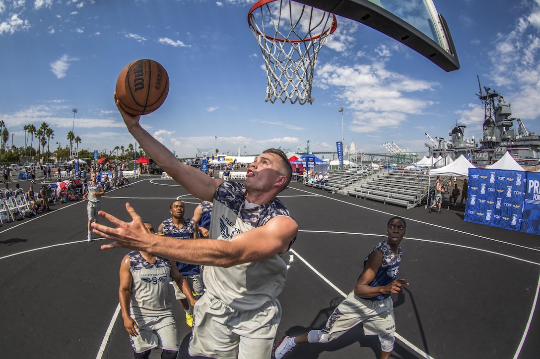 A sailor performs a layup at a basketball tournament with a ship in the background.