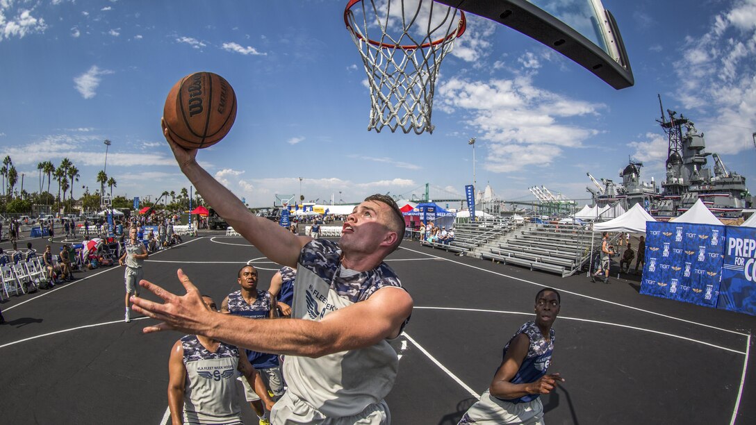 A sailor performs a layup at a basketball tournament with a ship in the background.