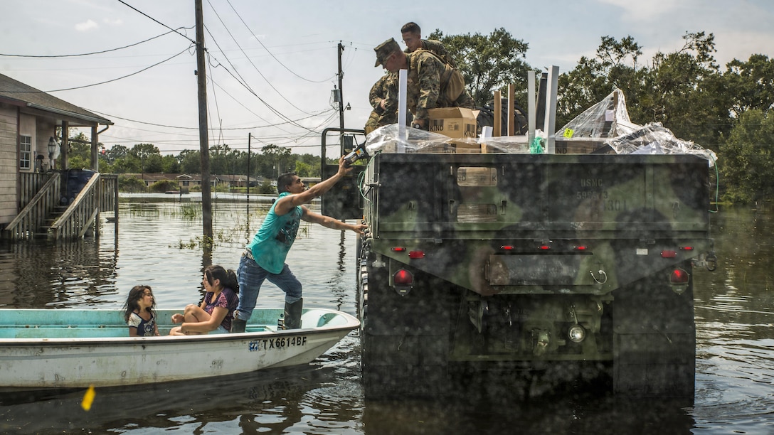 Marines in a military vehicle in a flooded area hand supplies to a man in a boat with two girls.