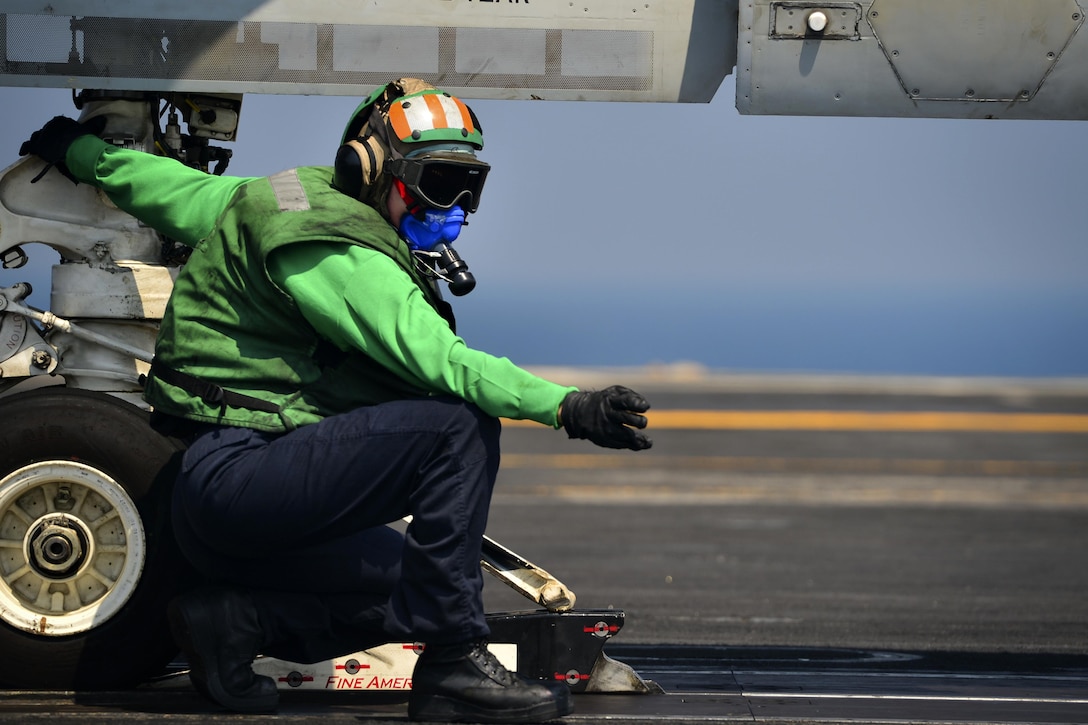 A sailor wearing heat gear makes signals while holding onto the wheel of an aircraft.