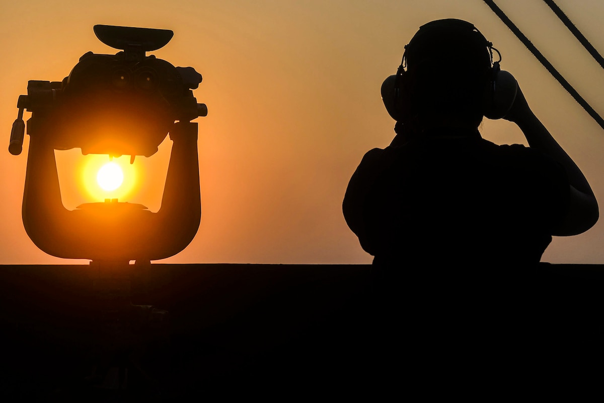 A sailor, shown from the back in silhouette, looks out from the deck of an aircraft carrier.