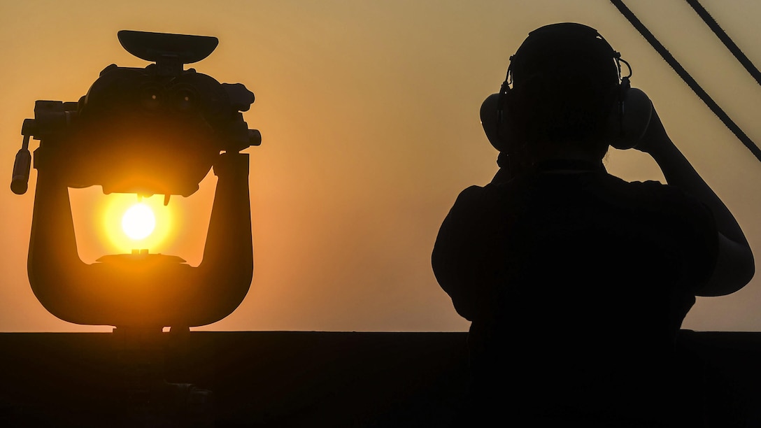 A sailor, shown from the back in silhouette, looks out from the deck of an aircraft carrier.