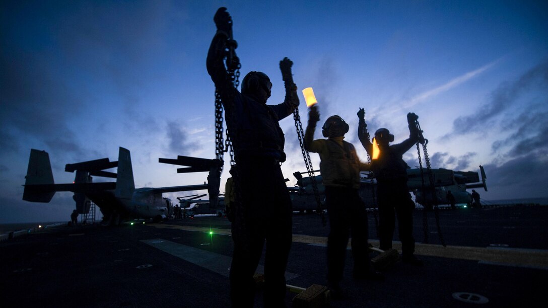 Three silhouetted figures hold up chains on a ship's flight deck.