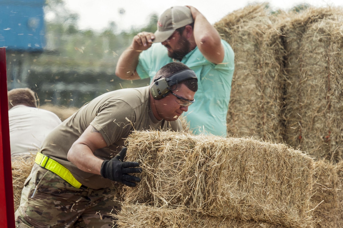 A service member lifts a bale of hay as small pieces of hay fly around him.