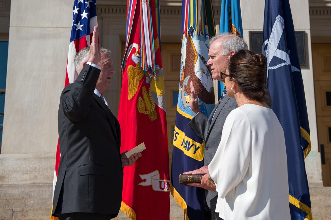 Defense Secretary Jim Mattis holds a book and his right hand in the air as he stands across from the incoming Secretary of the Navy.