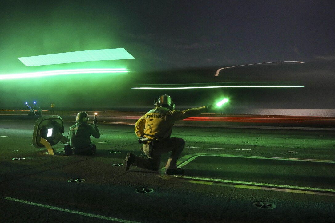 An aircraft lands on the flight deck of a ship at night as green lights flare.