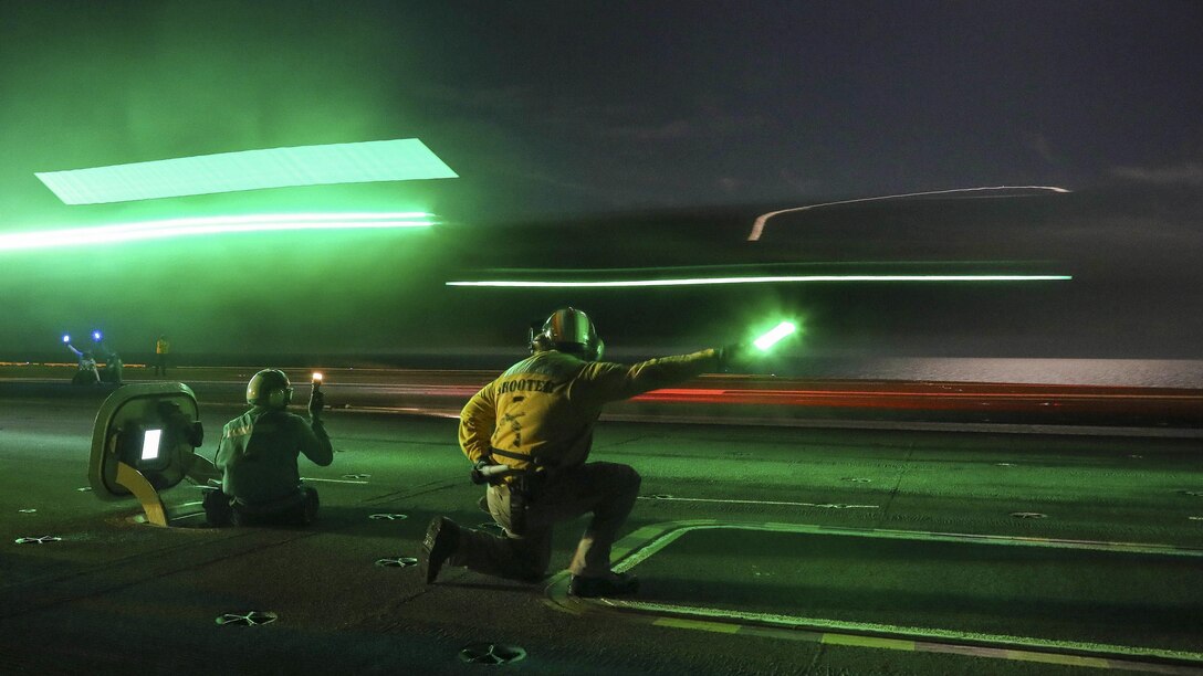An aircraft lands on the flight deck of a ship at night as green lights flare.