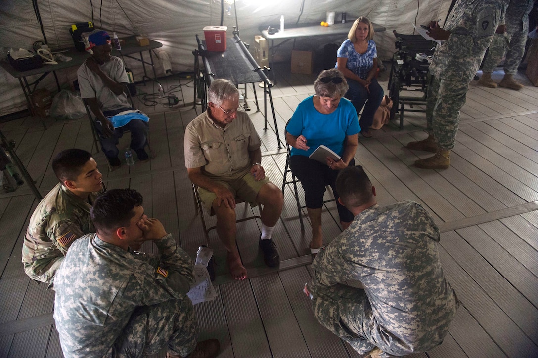Two civilians sit in chairs and talk to soldiers.